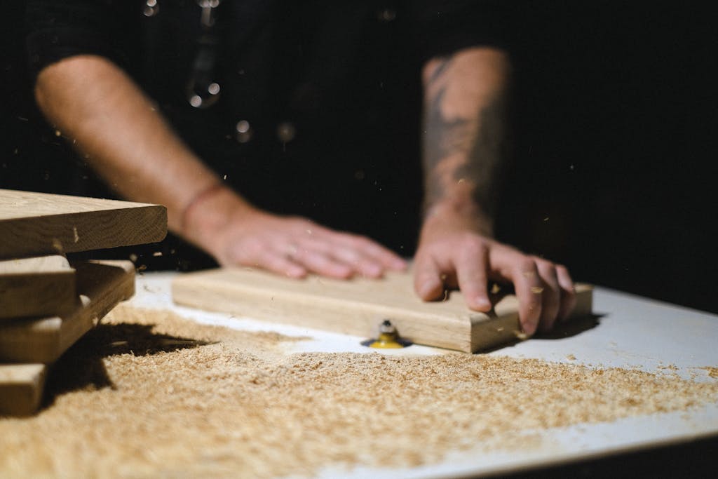 Unrecognizable male master standing at router table while shaping wooden plank at workbench with shavings in dark carpentry on blurred background